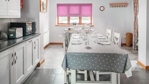 Uma área de jantar com mesa posta para quatro, cadeiras brancas, janela com cortina rosa e um relógio na parede.