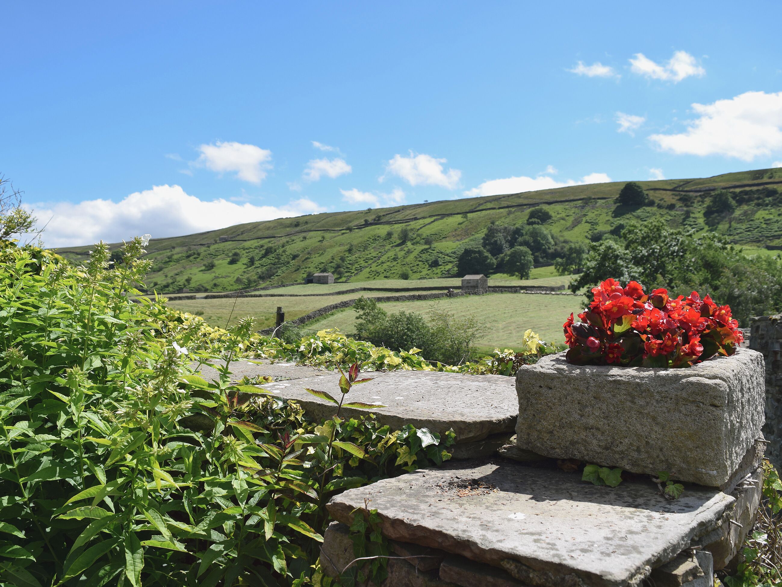 A stone bench with a flower arrangement in a rural landscape with rolling hills and a clear sky.