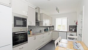 Eine moderne Küche mit weißen Schränken, einer Mikrowelle, einem Backofen und einem Geschirrspüler. Ein Fenster sorgt für natürliches Licht, und ein kleiner Tisch mit Stühlen steht bereit.