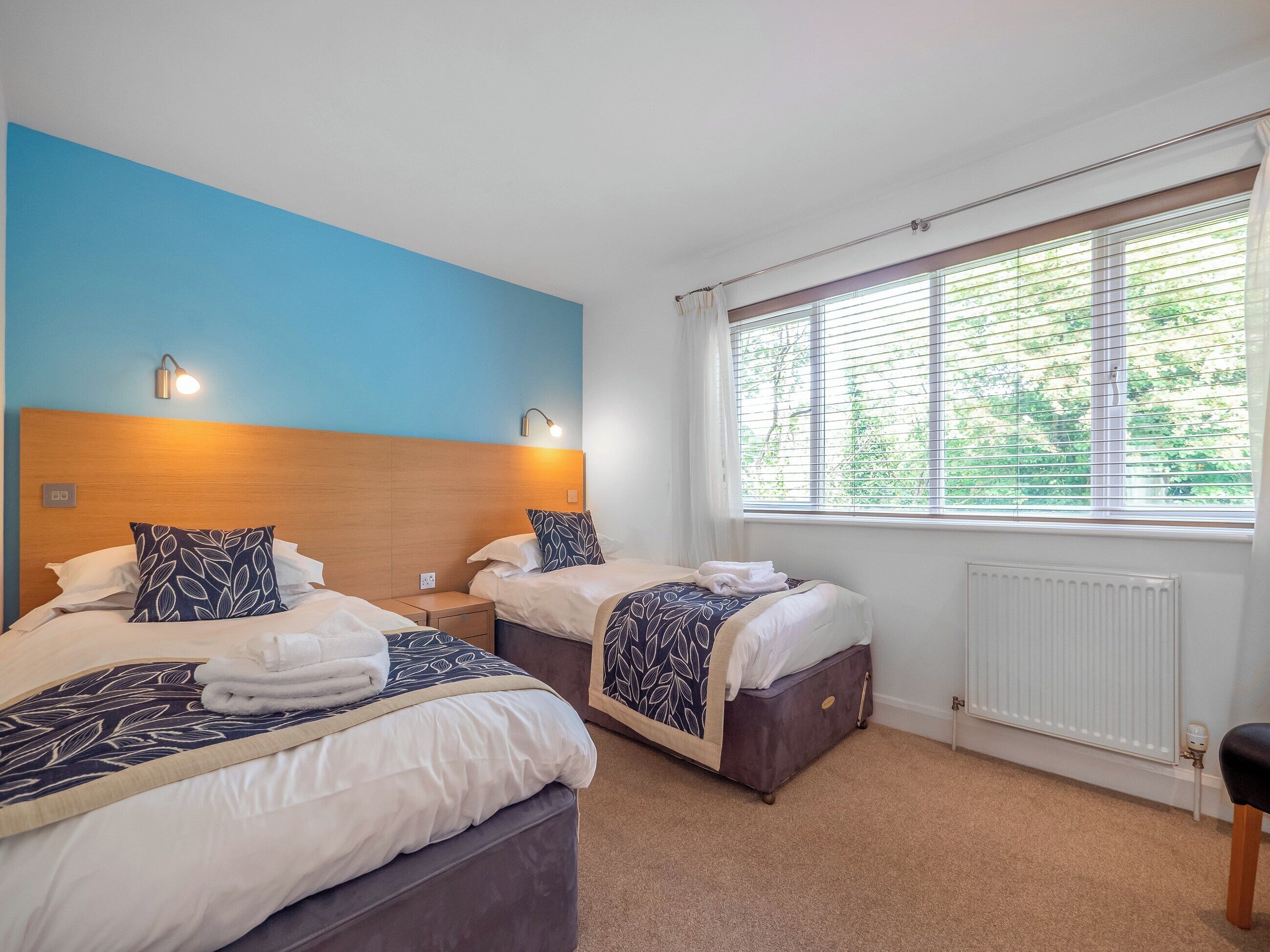A hotel room with two beds, a large window with blinds, a radiator, and a nightstand with a lamp.