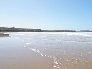 Uma praia com ondas suaves, céu claro e colinas ao longe.