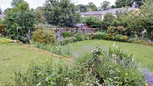 Ein Garten mit verschiedenen Pflanzen und Blumen, eine Steinmauer und ein Haus im Hintergrund.