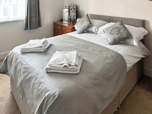 Cama bem arrumada com toalhas dobradas, mesa de cabeceira com um vaso e um espelho, e janela com cortinas.