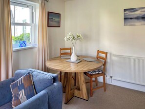 A room with a round wooden table, two chairs, a blue sofa, a white vase with flowers, a framed picture on the wall, and a window with curtains. - A ground floor apartment with level access, in Keswick. (Keswick)