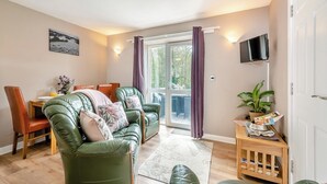 Ein gemütliches Wohnzimmer mit einem grünen Ledersofa, einem Holzesstisch mit Stühlen, einem wandmontierten Flachbildfernseher und einer Glastür, die zu einem Garten führt.