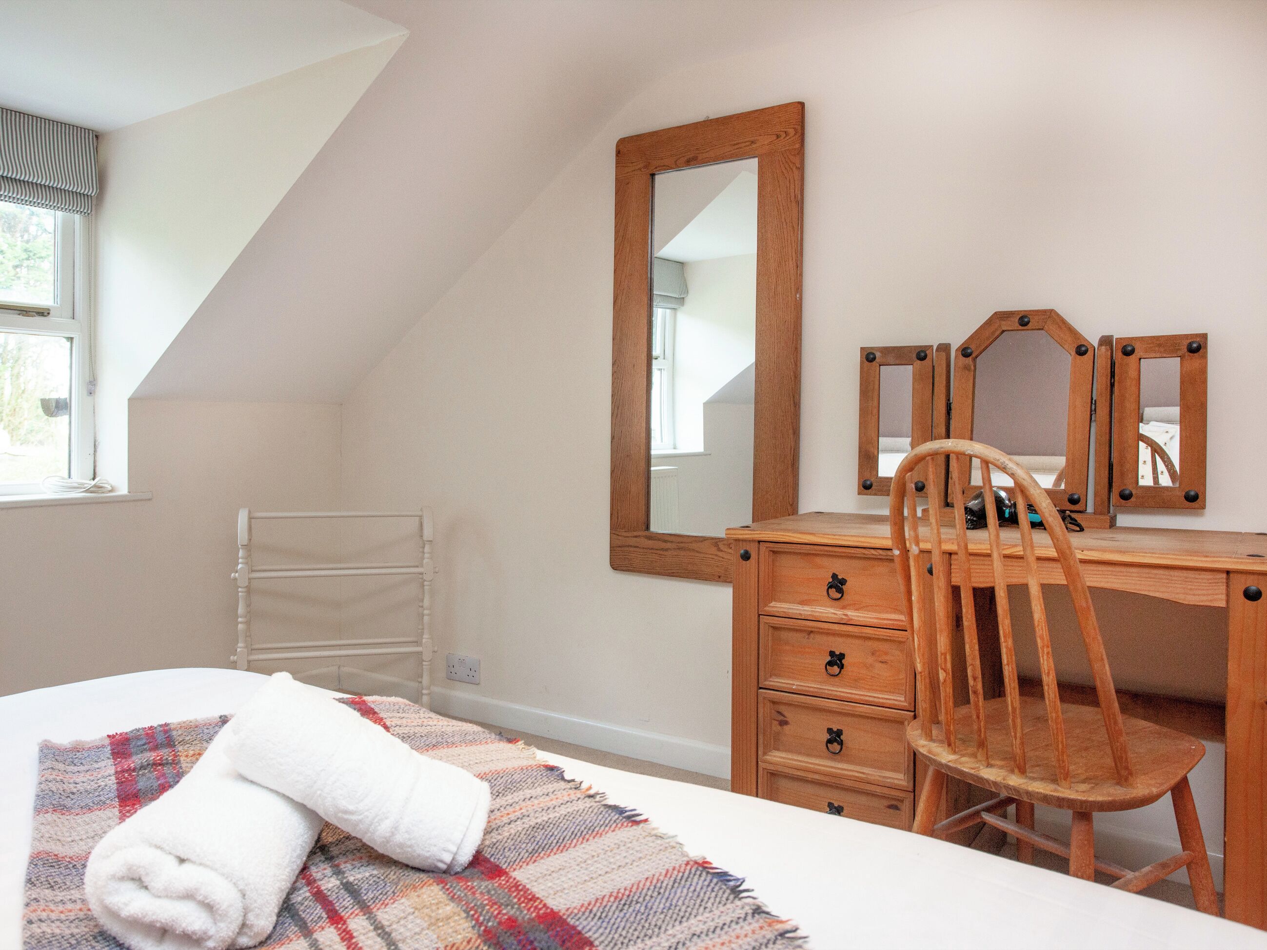 Quarto com cama, cómoda de madeira, cadeira, espelho grande e janela com cortinas.