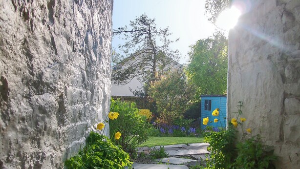 Un étroit sentier de pierre, bordé de murs de pierre rustiques, mène à un jardin où poussent des fleurs jaunes et se dresse une structure bleue.