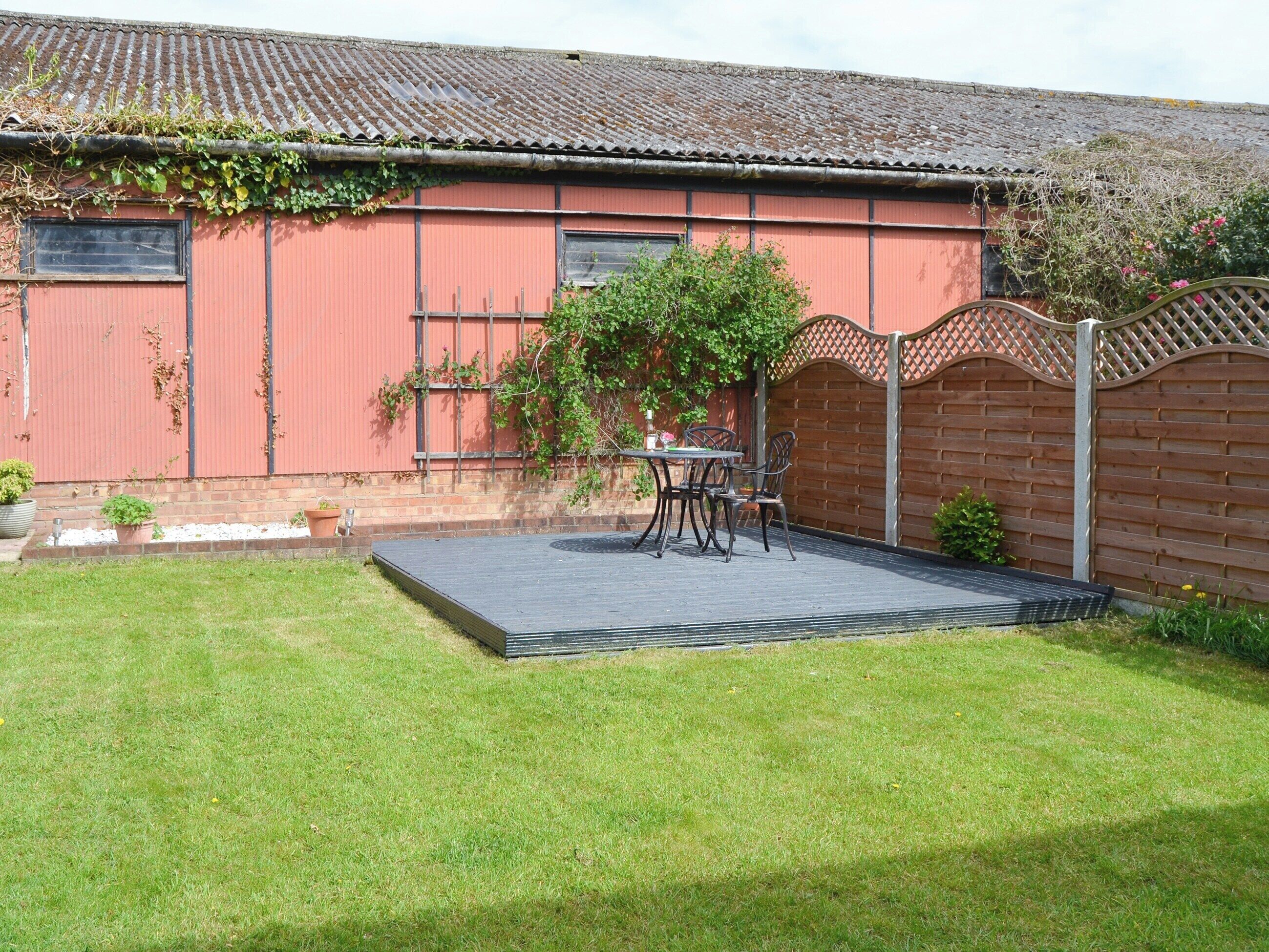 A red building with a tiled roof, a fenced garden, and a patio with a table and chairs.