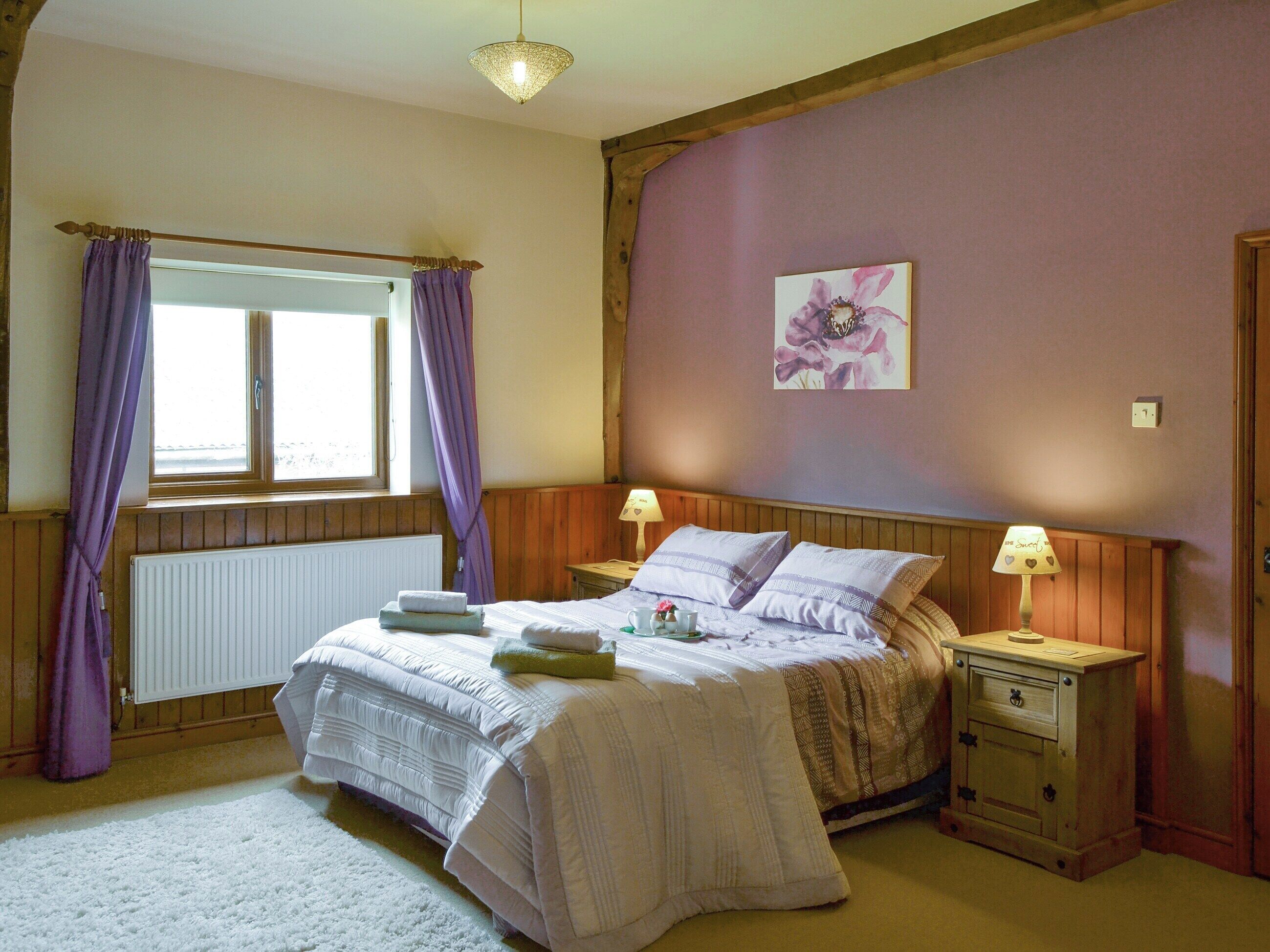 Une chambre à coucher avec un lit, deux tables de chevet, une fenêtre avec des rideaux violets, un radiateur et un tableau accroché au mur.