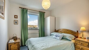 A bedroom with a bed, a nightstand, a dresser, a lamp, a window with curtains, and a radiator. - Sandringham Court Flat 4 (Swanage)