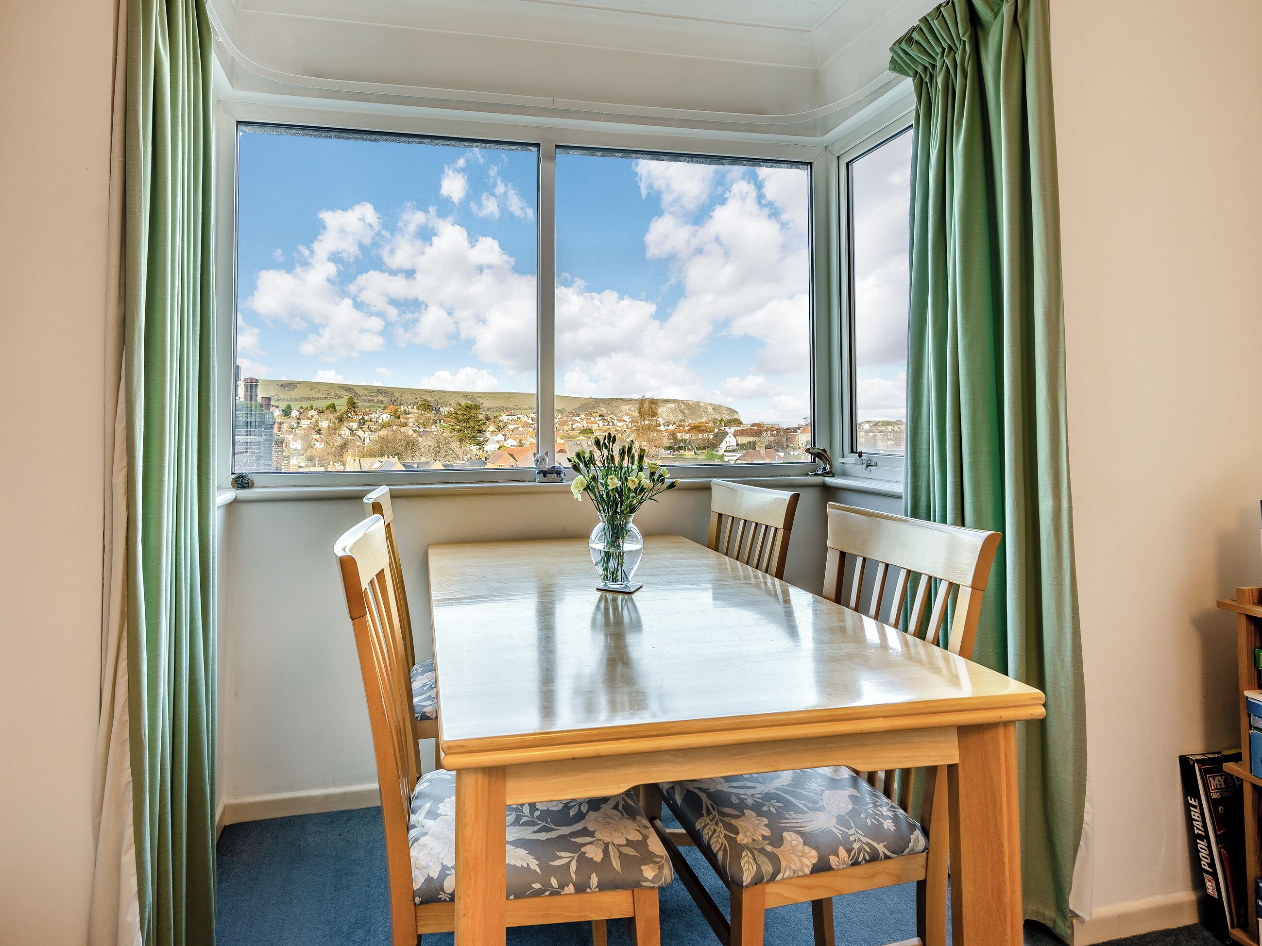 A dining area with a table and chairs, a vase with flowers, and a view of a town through large windows.