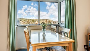 A dining area with a table and chairs, a vase with flowers, and a view of a town through large windows. - Sandringham Court Flat 4 (Swanage)
