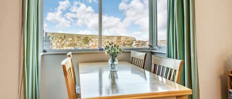 Uma área de refeições com uma mesa e cadeiras, um vaso com flores e vista para uma cidade através de grandes janelas.