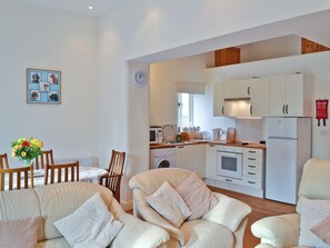 A modern kitchen with white cabinets, a microwave, and a refrigerator. There is a dining area with a table and chairs, and a living room with a sofa and cushions.