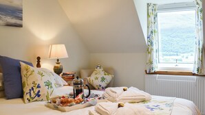 Ein Schlafzimmer mit einem Bett, Kissen, einem Nachttisch mit Lampe, einem Fenster mit Vorhängen und Blick ins Freie.