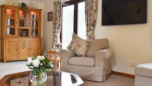 Ein Wohnzimmer mit einem beigen Sofa, einer Holzkabine mit Glastüren, einem flachbildfernseher an der Wand und einem Fenster mit blumigen Vorhängen.