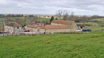 A stone building with a tiled roof, surrounded by a fenced field and a few cars parked nearby.