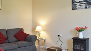 Un salon avec un canapé gris, une petite table avec une lampe, un vase rouge contenant des fleurs et un poêle à bois noir.