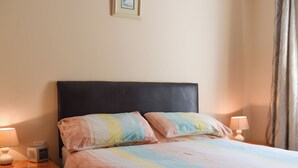 Un lit recouvert d’une couvre-lit à rayures, deux lampes de chevet, une table de chevet avec une lampe et un petit appareil électronique, et un tableau encadré accroché au mur.