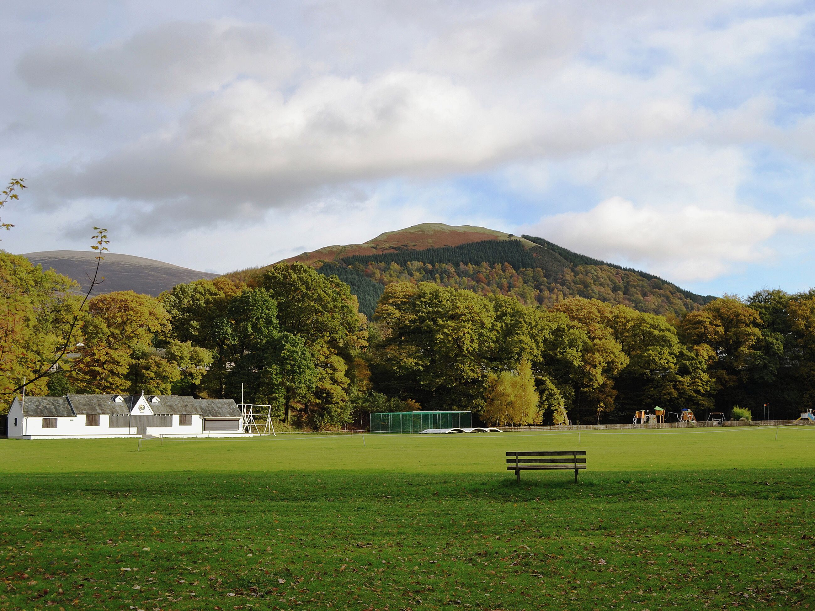 A grassy field with a bench, a white building with a sloped roof, and a forested hill in the background.