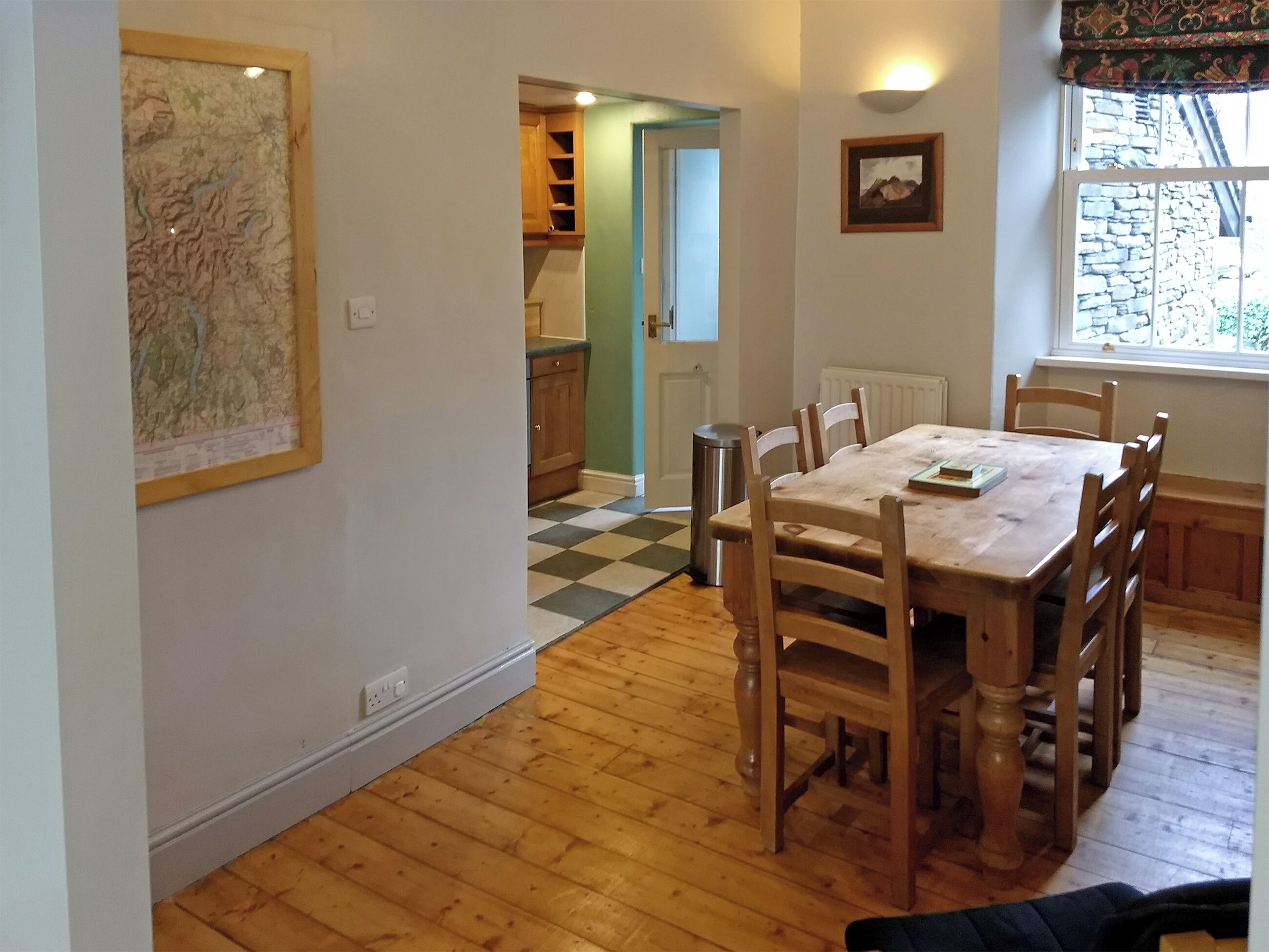 Uma área de refeições com mesa e cadeiras de madeira, mapa na parede, janela com vista e porta que dá para outro cômodo.