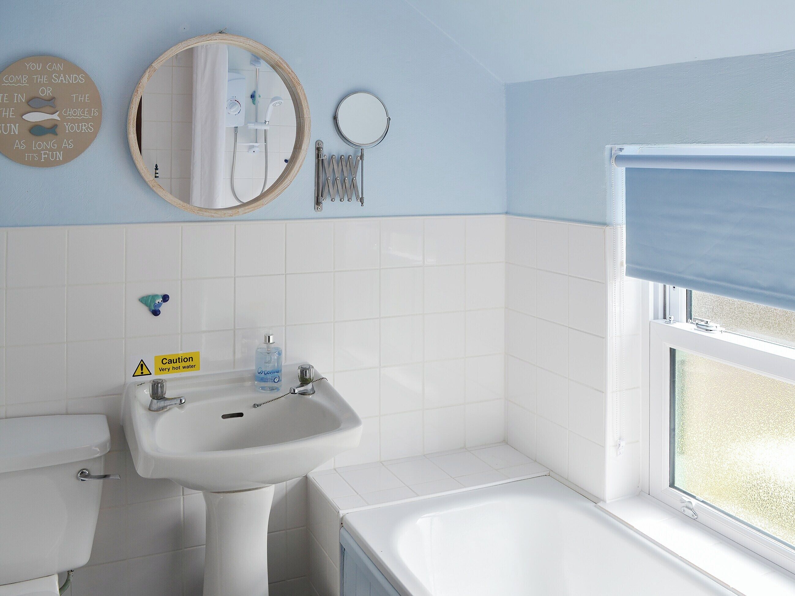 Une salle de bain équipée d’un lavabo, de toilettes et d’une baignoire. On y trouve une fenêtre avec un store, un miroir et un panneau d’avertissement indiquant que l’eau est chaude.