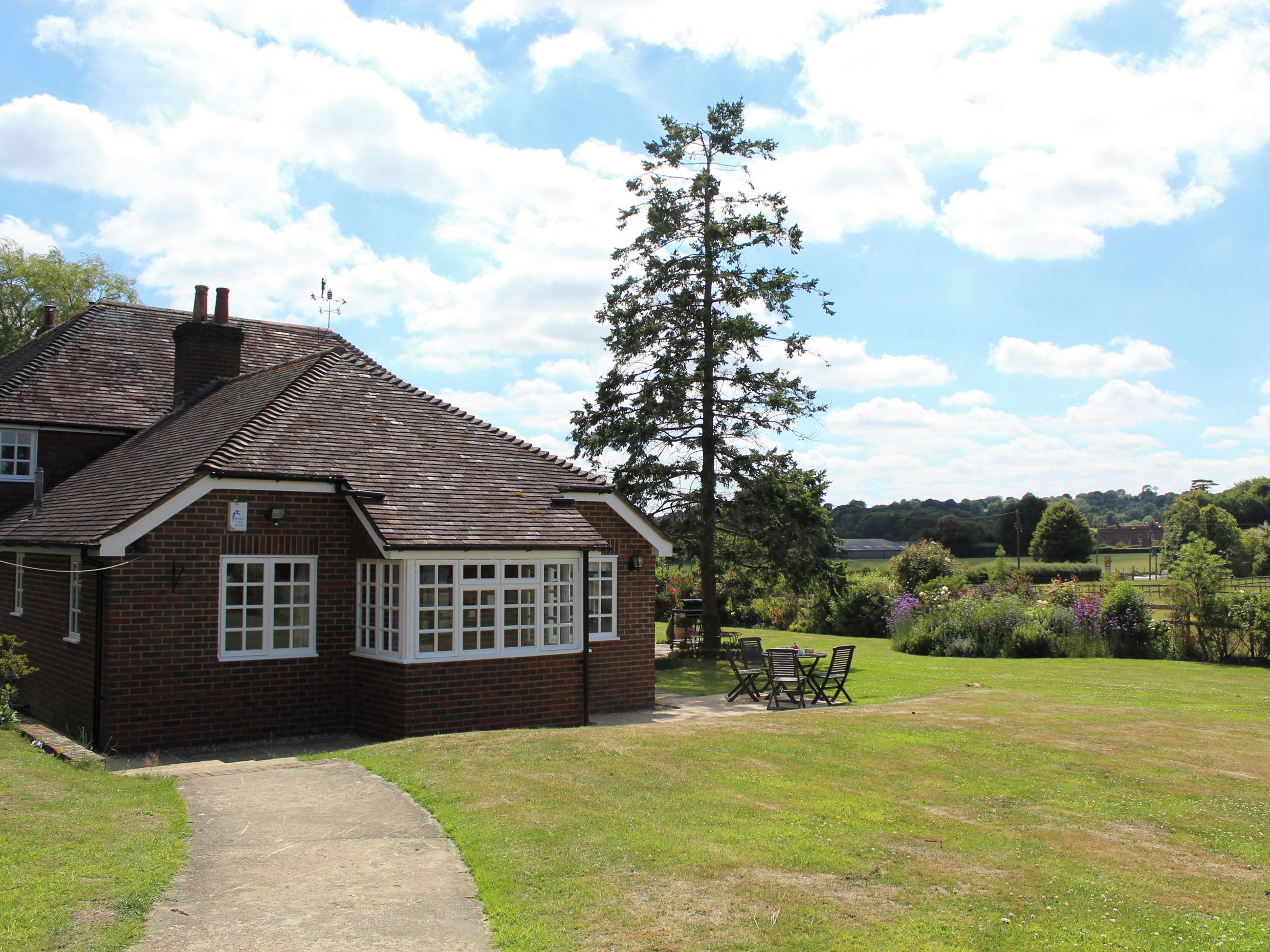 A brick house with a tiled roof, a large window, and a lawn with a table and chairs.