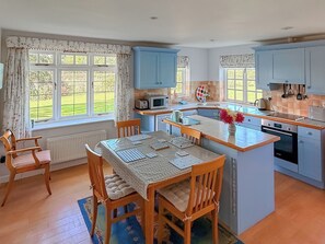 A kitchen with a dining area, wooden chairs, a table with a patterned tablecloth, and a view of a garden through the windows. - Castle House Cottage (Horton, near Wimborne)