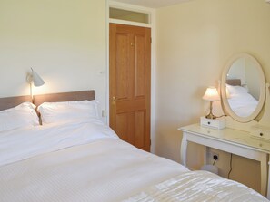 Een net opgemaakt bed met witte lakens, een bedlampje, een spiegel en een houten deur in een kamer met crèmekleurige muren.