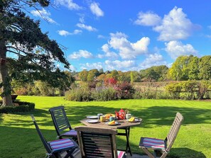 An outdoor dining area with a table set for two, surrounded by lush greenery and a clear blue sky. - Castle House Cottage (Horton, near Wimborne)