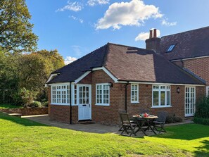 A two-story brick house with a pitched roof, a front porch, and a small garden area with a table and chairs. - Castle House Cottage (Horton, near Wimborne)