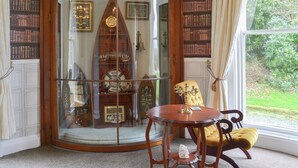 A room with a large arched window, a wooden table, a chair, and a bookshelf filled with books. - 2 bedroom accommodation in Liskeard (Liskeard)