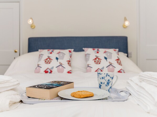 A neatly made bed with a blue headboard, white bedding, and two pillows with a red and white pattern. A tray with a book, a plate with a cookie, and a blue and white mug are placed on the bed.