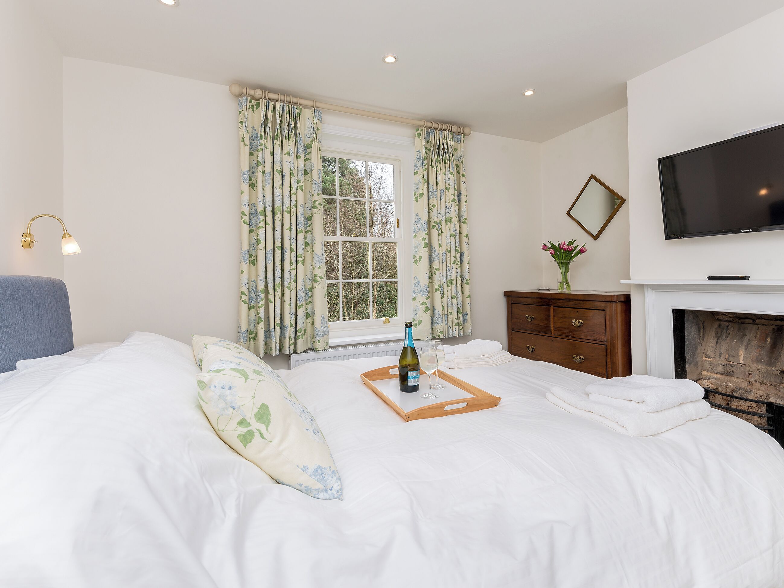 A bedroom with a bed, a bedside table with a bottle and glasses, a dresser, a TV, and a fireplace.