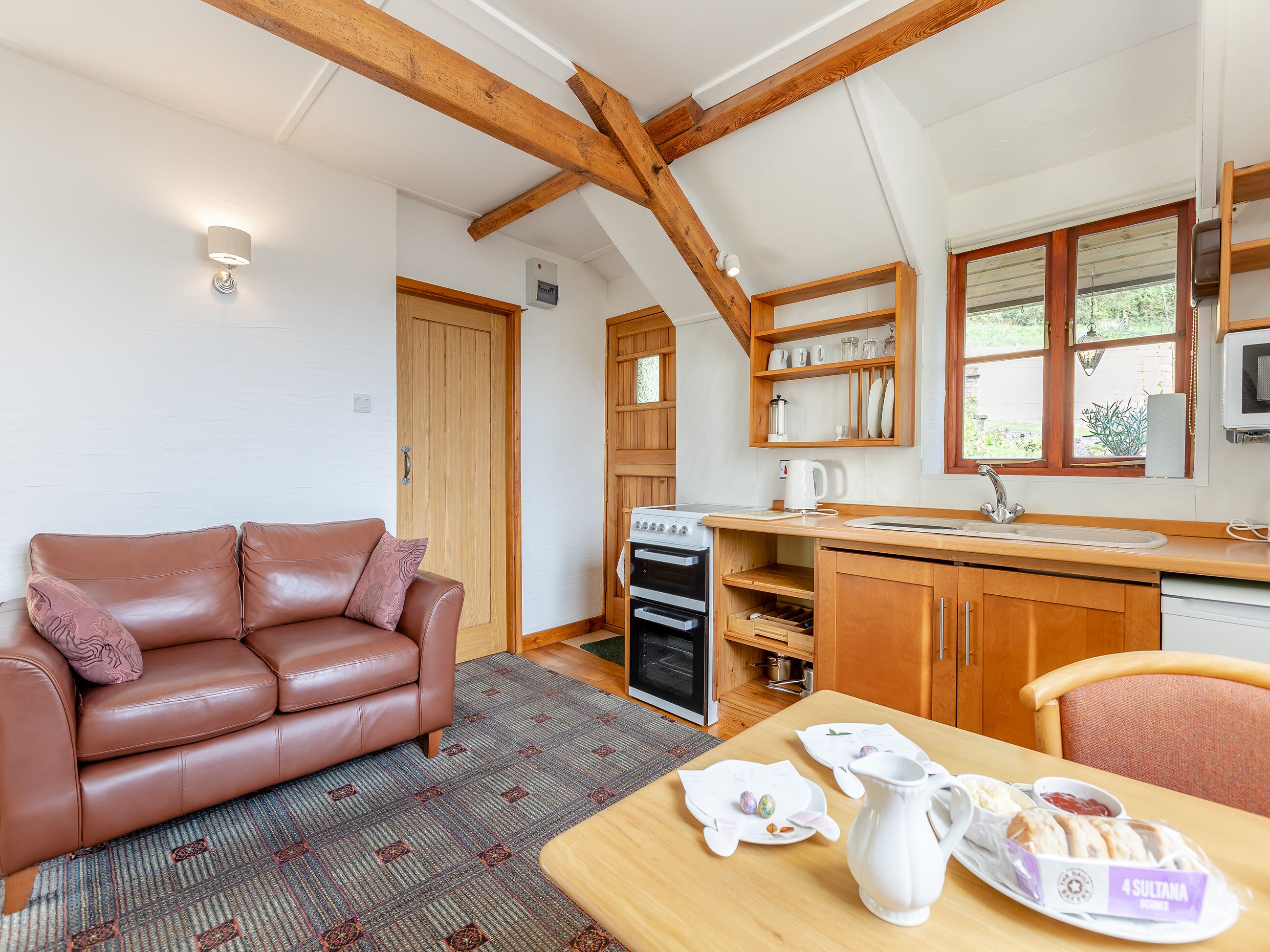 A cozy room with a brown leather sofa, a wooden dining table with a pitcher and plates, a kitchen with wooden cabinets and a stove, and a window with a view of greenery.