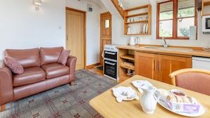 A cozy room with a brown leather sofa, a wooden dining table with a pitcher and plates, a kitchen with wooden cabinets and a stove, and a window with a view of greenery.