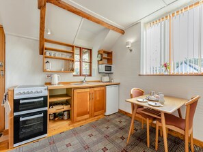 A compact kitchen with a stove, oven, and microwave. There is a wooden dining table with chairs, a window with blinds, and a small shelf with kitchenware. - 1 bedroom accommodation in Berrynarbor, near Ilfracombe (Berrynarbor, near Ilfracombe)