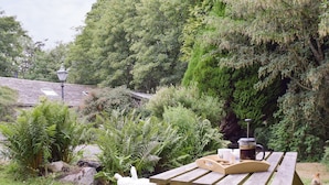 Une table de pique-nique en bois, avec une cafetière, des tasses et un plateau en bois, dans un jardin où poussent des fougères et où l’on aperçoit un oiseau blanc.