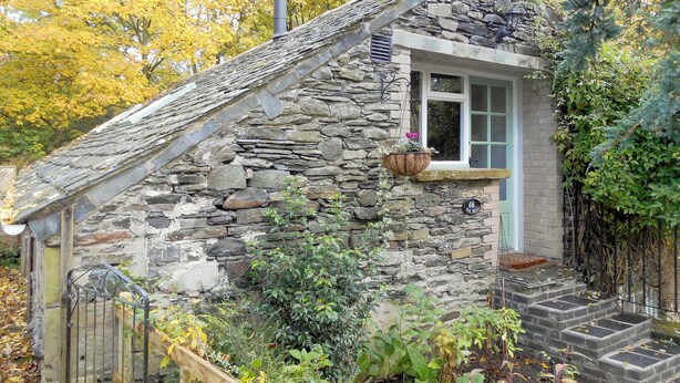 Une petite maison en pierre avec un toit à forte pente, une petite fenêtre et un escalier en pierre menant à l’entrée.