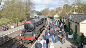 A vintage steam train at a station platform with passengers waiting.