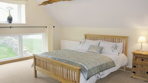 A bedroom with a wooden bed, a nightstand, a lamp, and a view of a grassy area through a window.
