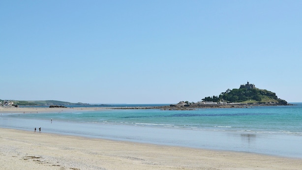 Ein Strand mit einem Felsen im Hintergrund, klarem Himmel und wenigen Personen, die am Ufer entlanggehen.