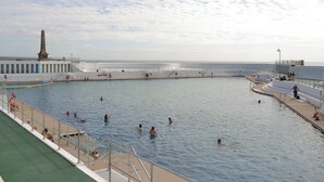 Ein großes öffentliches Schwimmbecken mit einem Denkmal im Hintergrund, umgeben von einem weißen Zaun und einer Promenade.