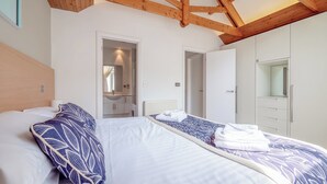 Un dormitorio con techo de madera, cama con ropa de cama blanca y azul, baño con espejo y lavamanos, y armario con espejo.