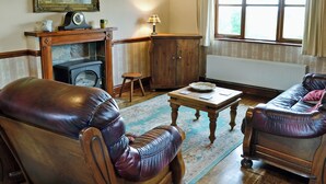 A traditional living room with leather armchairs, a wooden coffee table, a fireplace, a radiator, and a window with curtains. - 1 bedroom accommodation in Evershot (Evershot)