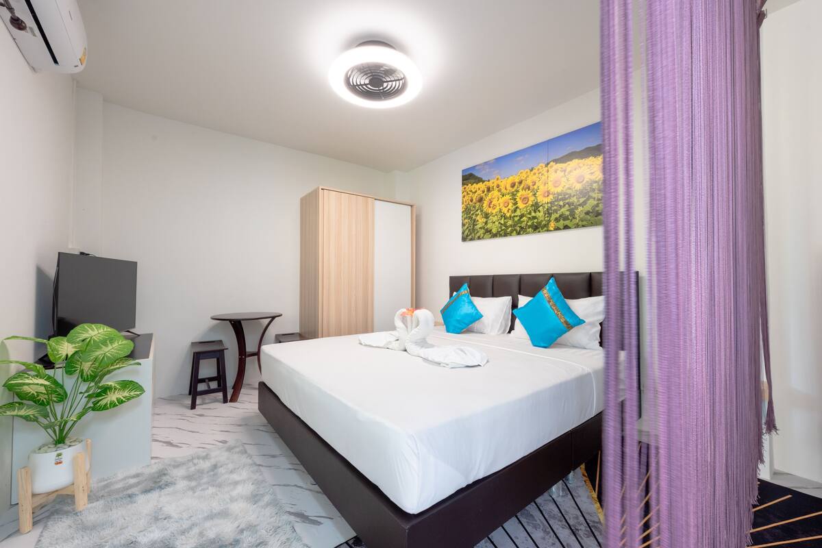 Habitación de hotel con cama, escritorio, televisor, planta y un cuadro en la pared.