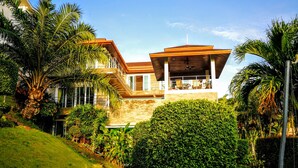 Una casa moderna con un balcón grande, rodeada de abundante vegetación y palmeras.