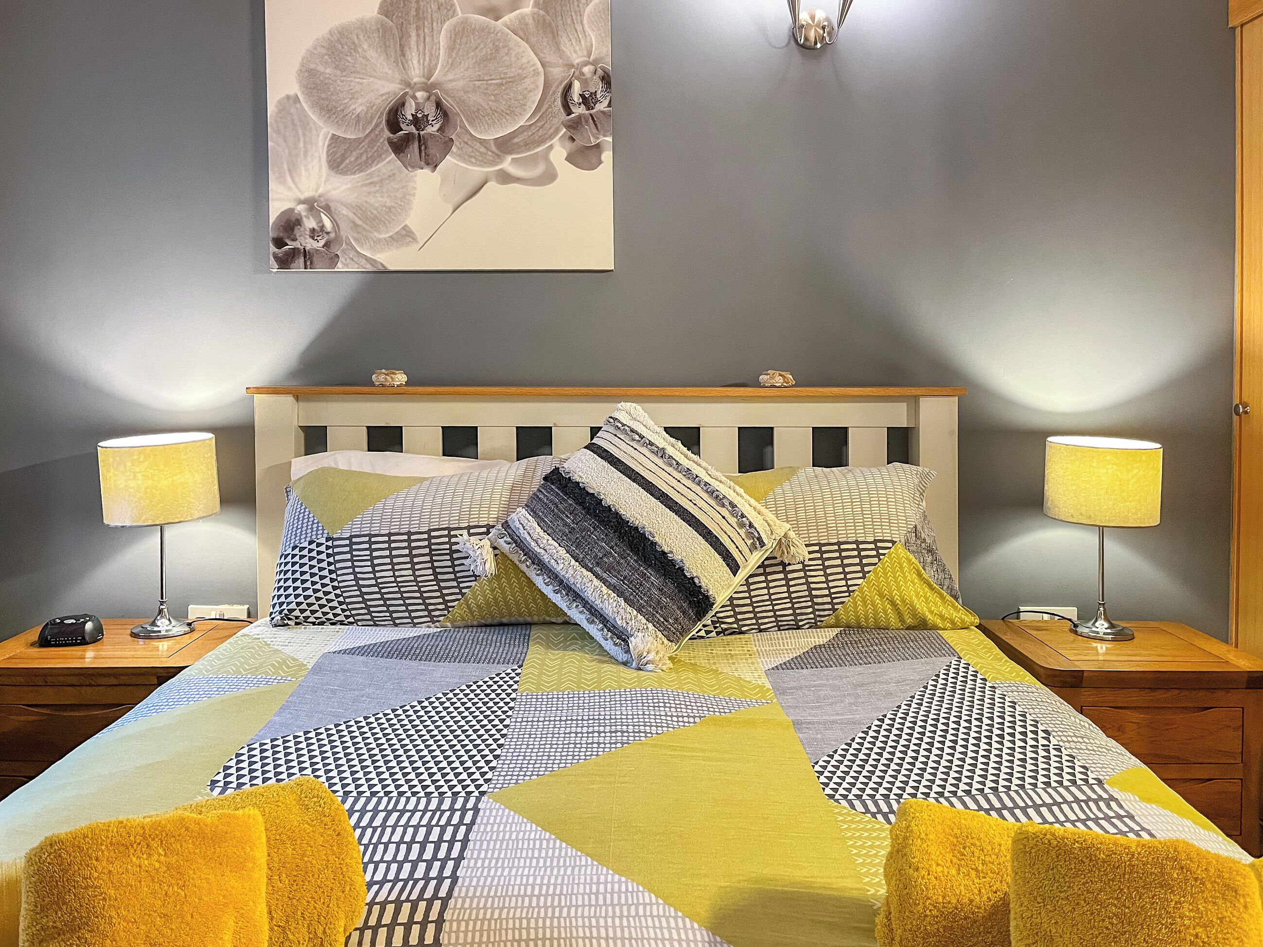 Una cama bien hecha con ropa de cama estampada, dos lámparas de noche y una obra de arte en la pared con orquídeas.