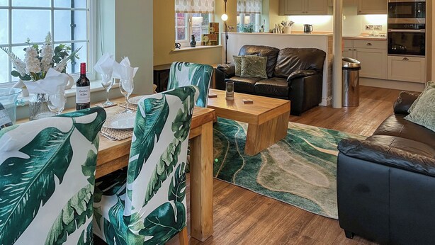 A cozy living room with a wooden coffee table, a sofa, and chairs with tropical leaf patterns. A dining area with a table and chairs is visible, along with a kitchen area with cabinets and appliances. - 2 bedroom accommodation in Filey (Filey)