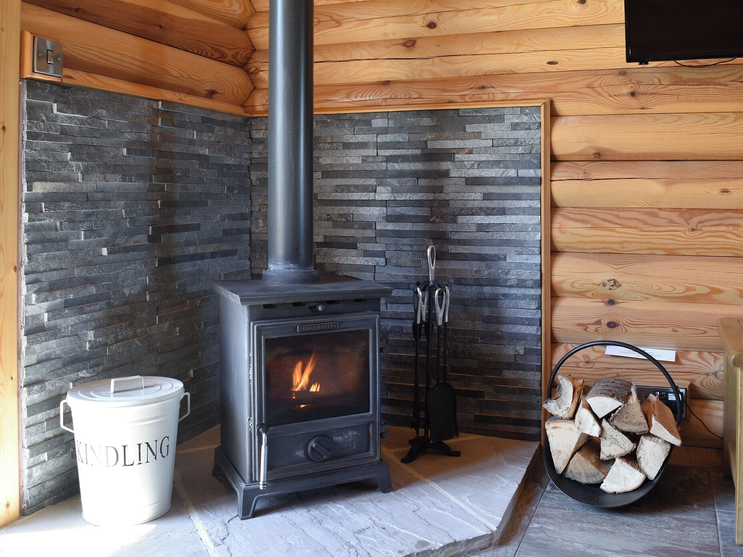 A wood-burning stove with a stone wall, a bucket labeled "KINDLING", and a pile of firewood.